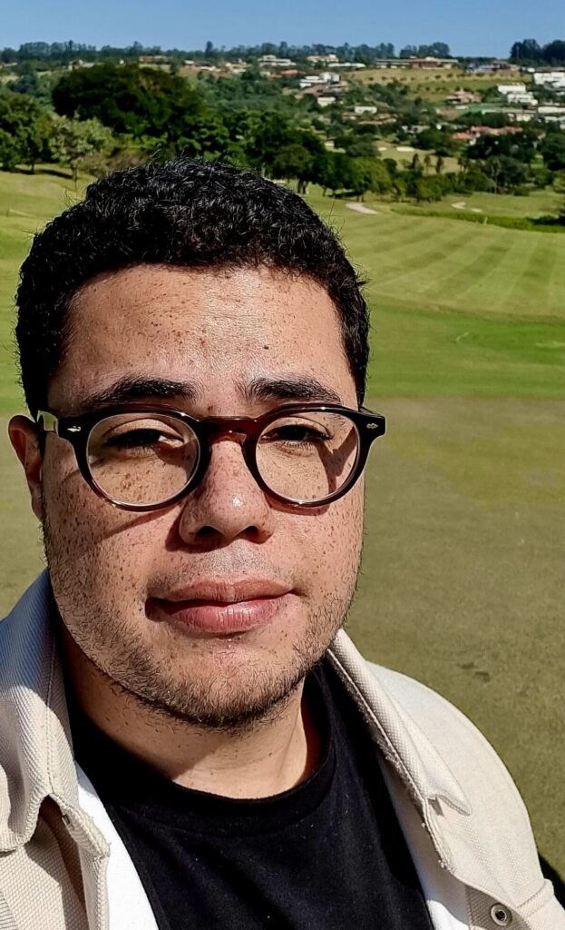 foto de tunai godoi, homem pardo com cabelos curtos castanhos escuros, óculos grossos de acetato, barba rala, sardas. Com uma aparencia amigável, de camiseta azul, camisa de piqué beige sobrepondo e posicionado em um campo de golfe extremamente verde e bem cuidado